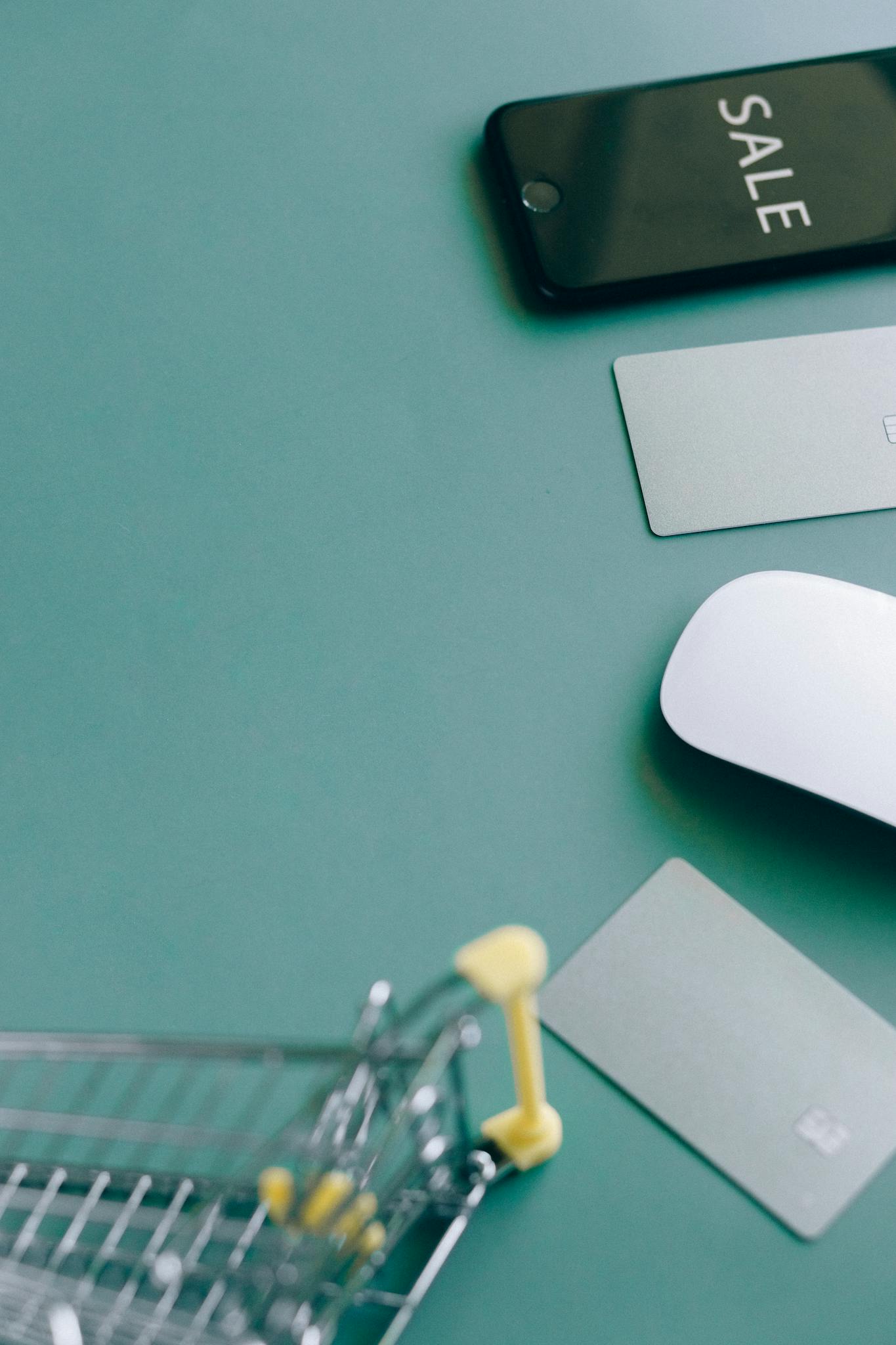 Flat lay of a shopping cart, phone with SALE display, and payment cards on a green background.